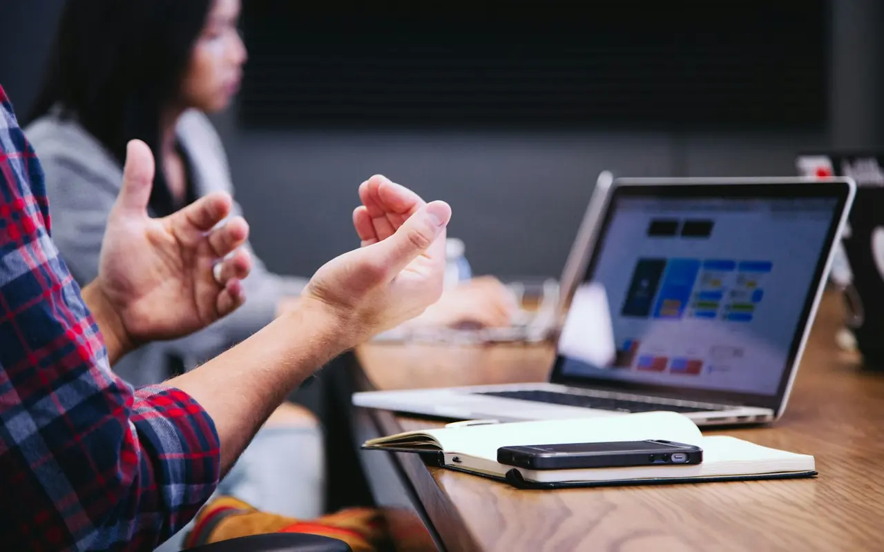 Due persone a un tavolo di lavoro: una spiega gesticolando, accanto un portatile aperto, un taccuino e un telefono