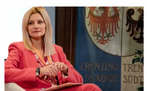 Eleonora Angelini seduta a un incontro pubblico, davanti allo stemma del Trentino-Alto Adige